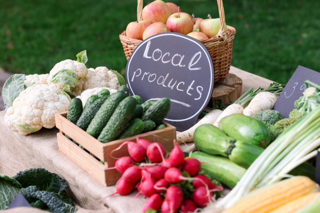 Korb mit Äpfeln und einer Tafel, auf der "Lokale Produkte" steht, umgeben von Blumenkohl, Gurken und Radieschen an einem Marktstand im Freien.