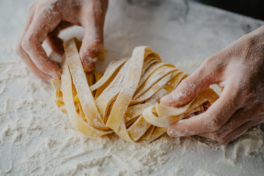 Close-up van meelbestoven handen die lange linten vers pasta vormen op een meelbedekt werkoppervlak.