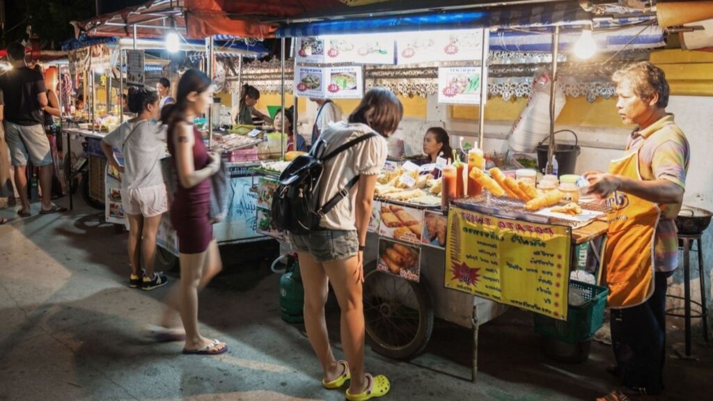 Night market food stall at dusk with customers lining up as a vendor grills skewers and displays a yellow menu board.