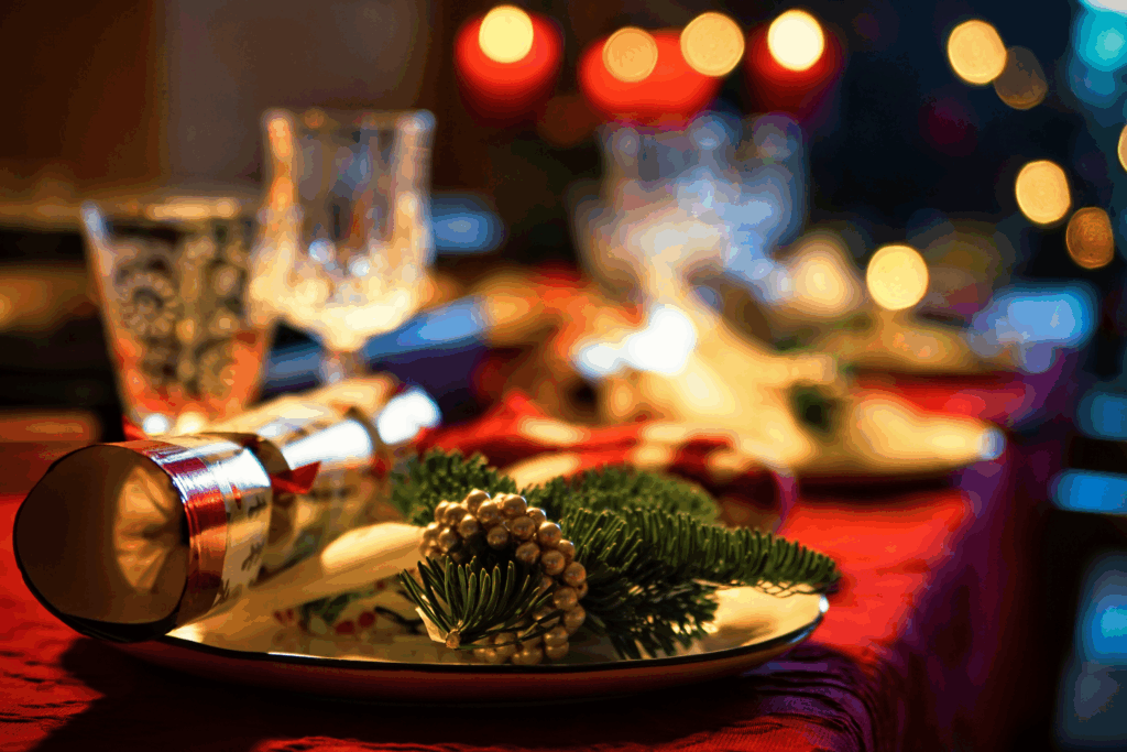 A holiday dinner table with a plate topped by pine sprigs and a decorative pinecone, blurred lights in the background.