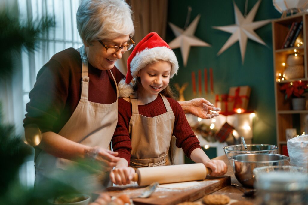 Grandmother and granddaughter rolling dough together in a festive kitchen, wearing aprons and Santa hat, smiling as they bake cookies.