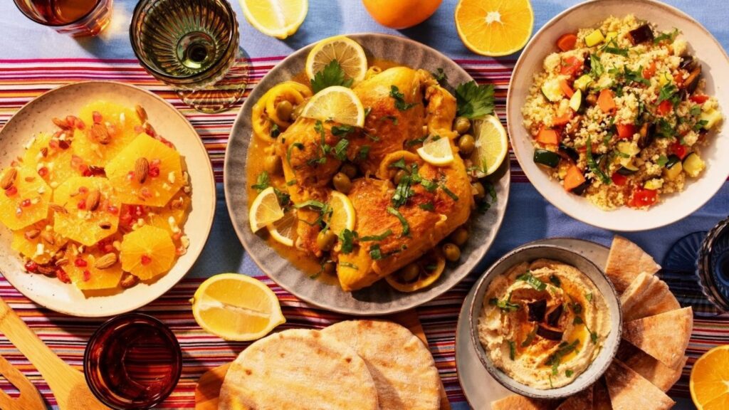 Vue de dessus d'un plateau coloré : poulet rôti garni de citron, segments d'orange avec grenade, salade de quinoa, houmous avec pain pita, et tranches de citron sur une nappe rayée.