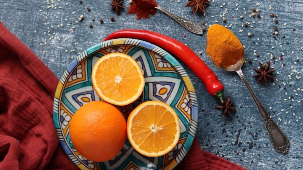Teller mit geschnittenen Orangen und einer ganzen Orange, einer roten Chilischote und Gewürzen auf einer blauen rustikalen Oberfläche. Dekorative, farbenfrohe kulinarische Szene.