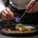A pastry chef carefully plating an ube purple yam dessert with deep violet cream and fresh garnish in a professional kitchen