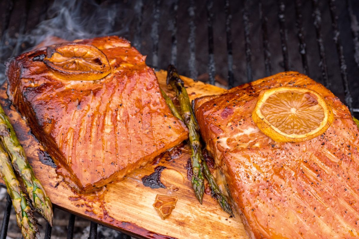 A cook brushing a miso marinade onto salmon before placing it among other grilled mains on a cedar plank