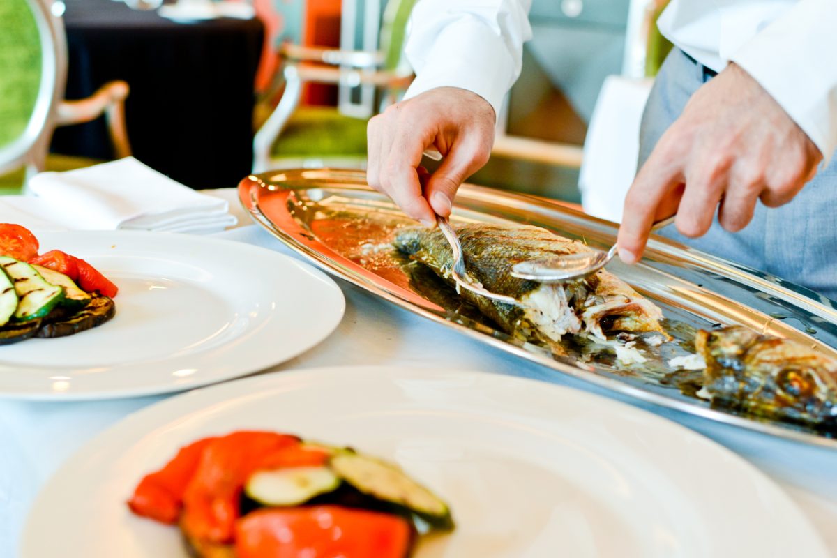 A woman presenting a platter of grilled mains including whole sea bass and prawns to guests at a summer outdoor table