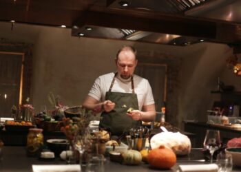 Chef in a white shirt and green apron plates food at a busy kitchen counter, surrounded by vegetables and bowls.