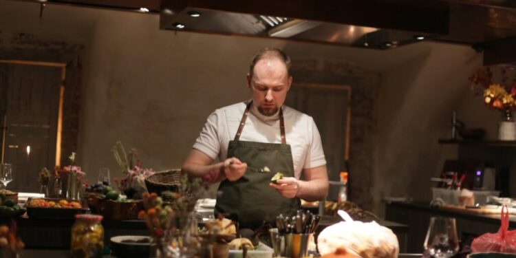 Chef in a white shirt and green apron plates food at a busy kitchen counter, surrounded by vegetables and bowls.