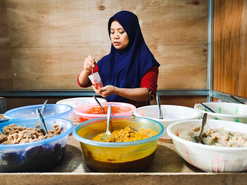Woman in a blue hijab scoops red sauce into a cup in front of several large bowls of colorful ingredients.