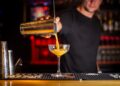 A smiling bartender pours a classic Aperol spritz cocktail into a large wine glass filled with ice and an orange garnish.