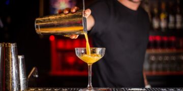 A smiling bartender pours a classic Aperol spritz cocktail into a large wine glass filled with ice and an orange garnish.