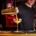 bartender-pouring-orange-spritz-glass-unsplash - GUSTOwebzine Ein lächelnder Barkeeper schenkt einen klassischen Aperol Spritz in ein großes Weinglas mit Eis und einer Orangenscheibe ein.