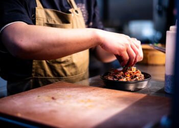 A chef carefully assembles a balanced grain bowl with layered grains, vegetables, and fresh herbs on a wooden counter