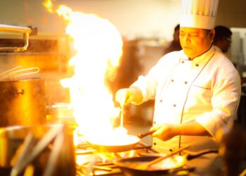 A chef holds tongs over an open flame while charring summer peppers on a gas stovetop in a professional kitchen.