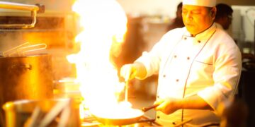 A chef holds tongs over an open flame while charring summer peppers on a gas stovetop in a professional kitchen.
