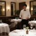 Fine Dining Economics Michelin Restaurant Dubai 2026 - GUSTOwebzine A waiter in a white shirt and black bow tie stands at a table in an elegant restaurant, checking a menu or notepad.
