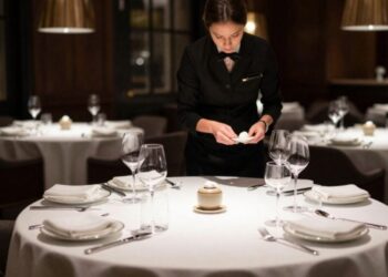Waiter in a black uniform arranges a formal dining table with wine glasses and white plates.