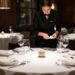 Waiter in a black uniform arranges a formal dining table with wine glasses and white plates.