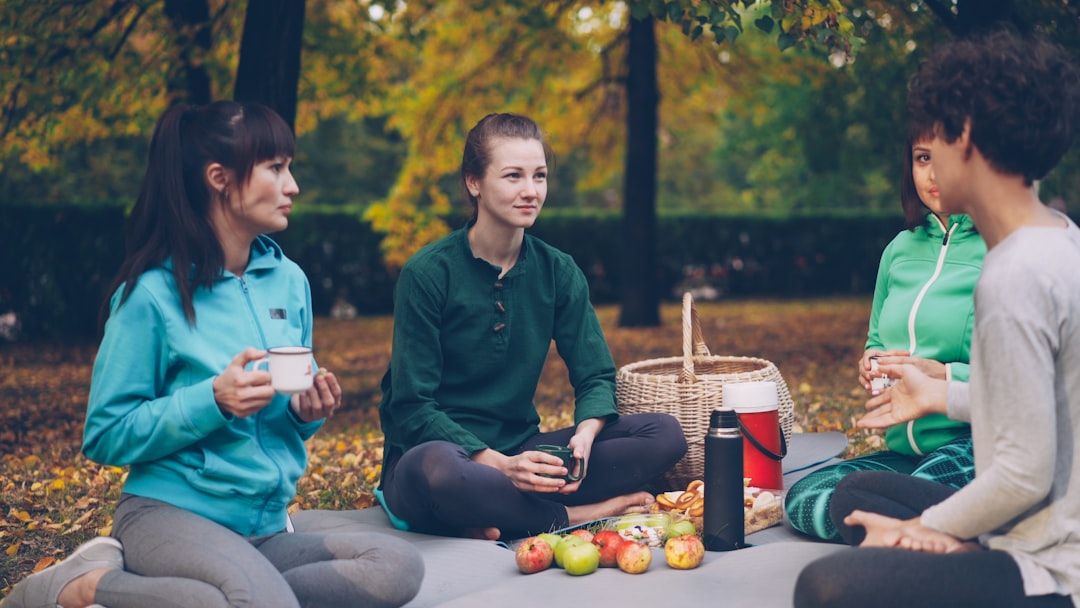 A group of friends sits on a blanket in a park, sharing plates of picnic recipes including  salads and pressed sandwiches.