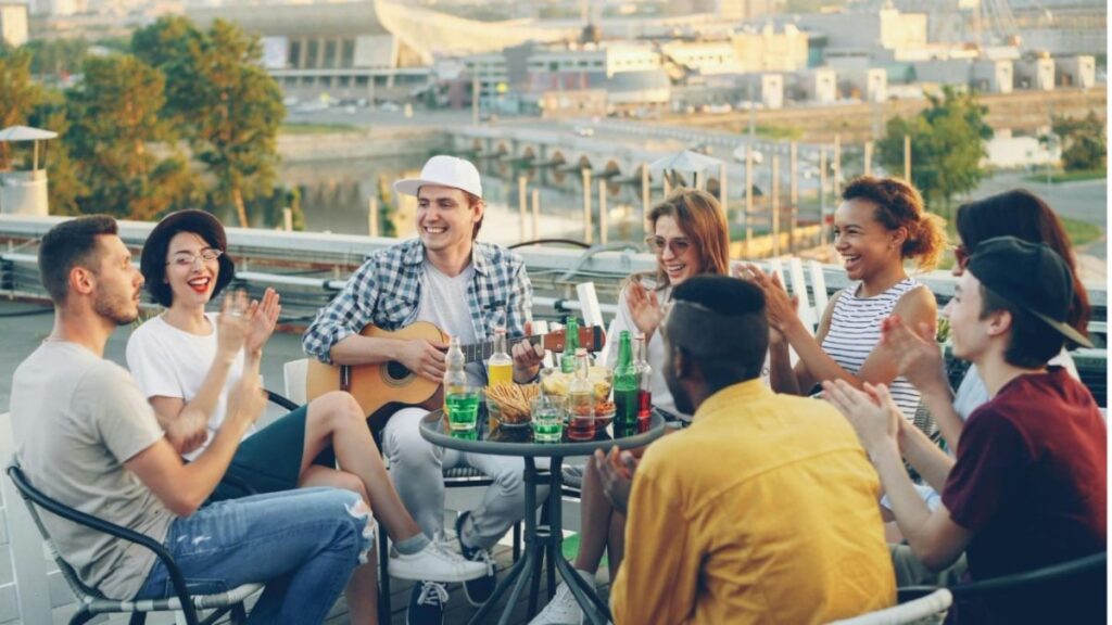 Group of friends sitting around a rooftop table, one person playing guitar and others clapping and laughing.