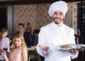 A chef steps out of a small restaurant kitchen to welcome loyal regulars at their table