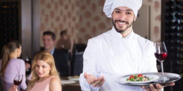 A chef steps out of a small restaurant kitchen to welcome loyal regulars at their table