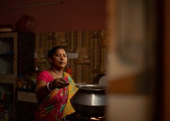 Indian woman in a colorful sari cooking over a metal pot on a stove, reaching with a ladle in a dimly lit kitchen.