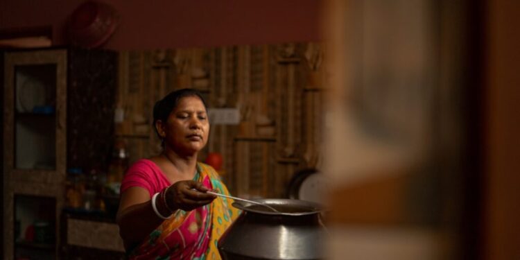 Gluten-free food India best dishes to try - GUSTOwebzine Indian woman in a colorful sari cooking over a metal pot on a stove, reaching with a ladle in a dimly lit kitchen.