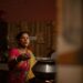 Gluten-free food India best dishes to try - GUSTOwebzine Indian woman in a colorful sari cooking over a metal pot on a stove, reaching with a ladle in a dimly lit kitchen.