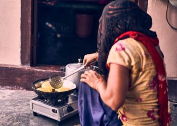 Woman in a colorful dress and red scarf frying round flatbreads on a pan over a portable gas stove outside a doorway.