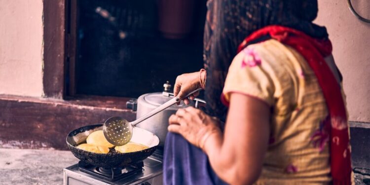 Gluten-free Indian breakfast with millet - GUSTOwebzine Woman in a colorful dress and red scarf frying round flatbreads on a pan over a portable gas stove outside a doorway.