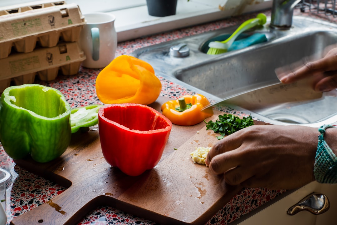 A home cook peels the blackened skin from charred summer peppers over a glass bowl on a wooden kitchen counter.