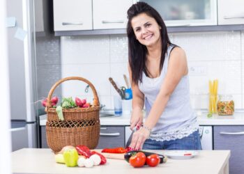 A woman carefully arranges containers of picnic recipes into a wicker basket on a kitchen, ready for a June outdoor outing.