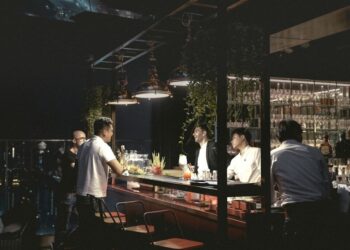 Nighttime rooftop bar with a bartender and a customer leaning on the illuminated counter, city lights in the background.