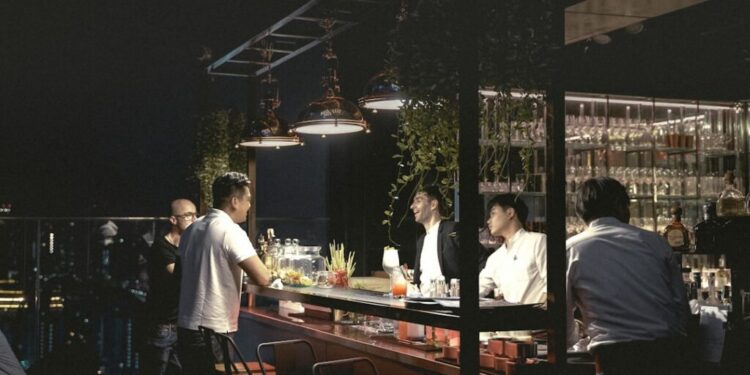 Bar en azotea nocturno con un cantinero y un cliente apoyado en la barra iluminada, luces de la ciudad de fondo.