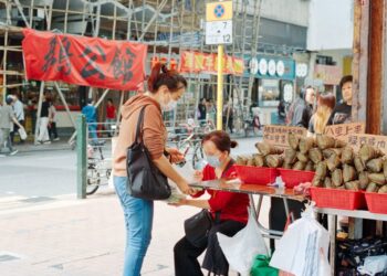 Woman in a mask buys sticky rice dumplings from a street vendor at an outdoor market stall with red baskets and stacked wraps on display.",