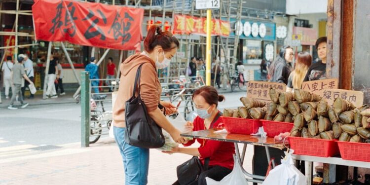 Woman in a mask buys sticky rice dumplings from a street vendor at an outdoor market stall with red baskets and stacked wraps on display.",
