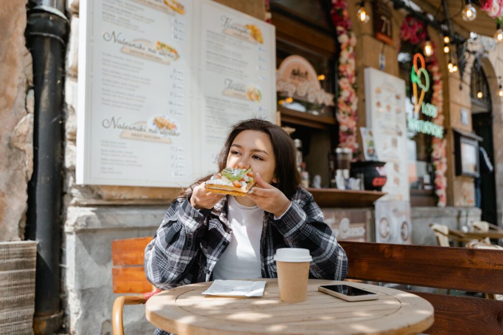 グルテンフリー パリ 8つの最高のビストロ - GUSTOwebzine 格子縞のシャツを着た女性が屋外のカフェでサンドイッチを食べている様子。テーブルの上にはコーヒーカップとスマートフォンがあり、彼女の後ろにはメニューがある。