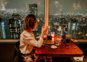 Woman seated at a candlelit restaurant table, toasting with a glass as city lights glow through a window behind her at night.