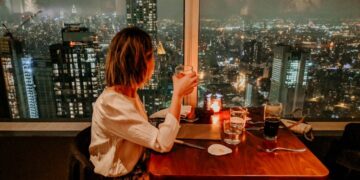 Femme assise à une table de restaurant à la lumière des bougies, trinquant avec un verre tandis que les lumières de la ville brillent par la fenêtre derrière elle la nuit.