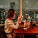 Gluten-free spots for summer dining NYC - GUSTOwebzine Woman seated at a candlelit restaurant table, toasting with a glass as city lights glow through a window behind her at night.