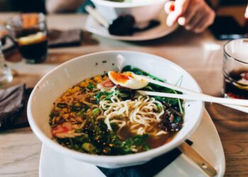 Bowl of ramen with noodles, corn, greens, and a half-boiled egg, chopsticks resting on top on a wooden table in a dining setting.