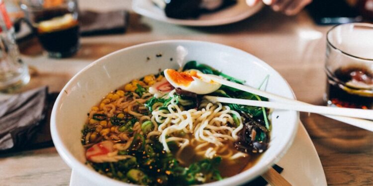 Gluten-free ramen Tokyo 7 great spots - GUSTOwebzine Bowl of ramen with noodles, corn, greens, and a half-boiled egg, chopsticks resting on top on a wooden table in a dining setting.
