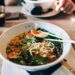 Bowl of ramen with noodles, corn, greens, and a half-boiled egg, chopsticks resting on top on a wooden table in a dining setting.