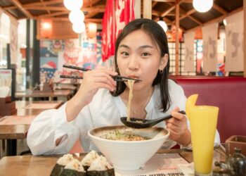 Young woman eats ramen in a casual restaurant, lifting noodles with chopsticks over a large bowl of noodles on the table.