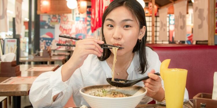 Young woman eats ramen in a casual restaurant, lifting noodles with chopsticks over a large bowl of noodles on the table.