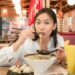 Flushing Chinatown your complete food guide - GUSTOwebzine Young woman eats ramen in a casual restaurant, lifting noodles with chopsticks over a large bowl of noodles on the table.