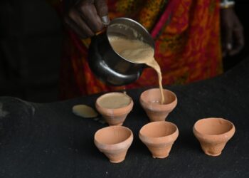 Person pours beige batter from a metal pitcher into five small terracotta cups on a dark surface.