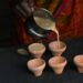 Person pours beige batter from a metal pitcher into five small terracotta cups on a dark surface.