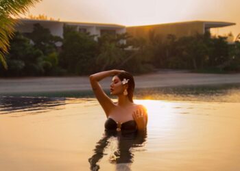 Woman in a black bikini standing chest-deep in a sunset-lit pool, hand resting on her head with a flower in her hair.